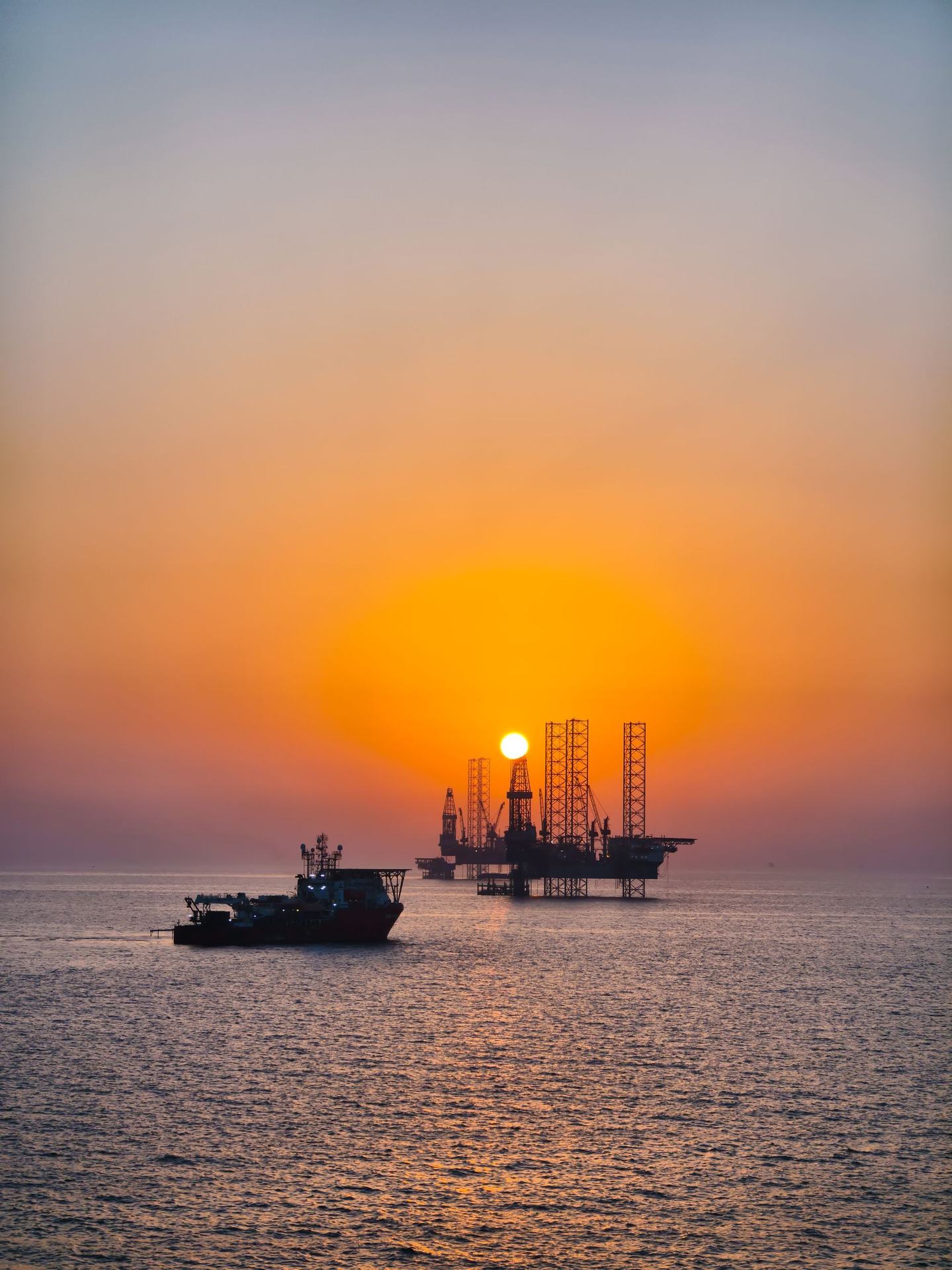 Offshore jack-up rigs and a supply vessel silhouetted at sunset